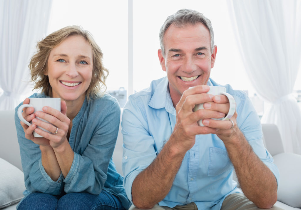 smiling middle-aged couple sitting on a couch and holding mugs, possibly containing coffee